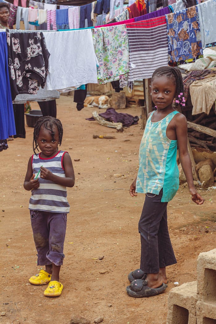 Two young girls standing in a village street in Abuja, surrounded by colorful hanging laundry.