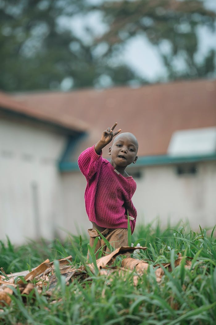 A child in a pink sweater makes a peace sign in a grassy rural setting, conveying innocence and hope.