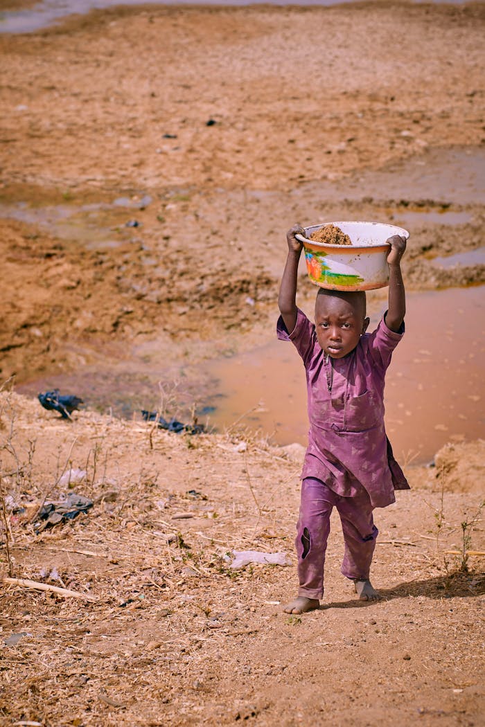 A young boy carries a bowl across a dry, rural landscape.