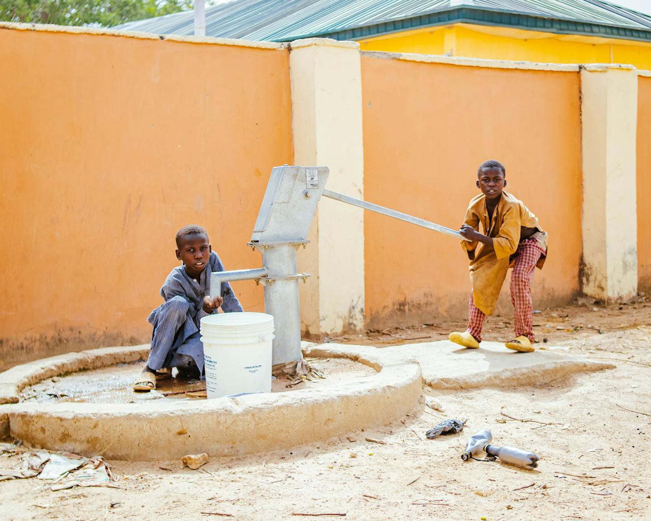 Two children use a manual pump to draw water into a bucket against an orange wall on a sunny day.